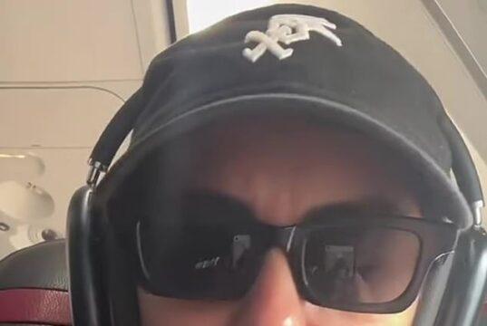 Sunglasses-wearing woman with headset on airplane, casual attire, serious expression.