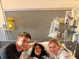Young girl in hospital bed with two smiling visitors, medical equipment in background.