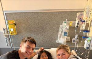 Young girl in hospital bed with two smiling visitors, medical equipment in background.