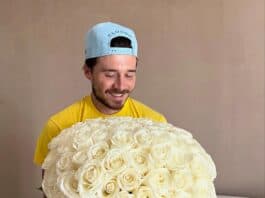 Vase filled with white roses held by smiling man wearing baseball cap.