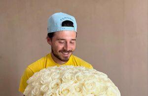 Vase filled with white roses held by smiling man wearing baseball cap.