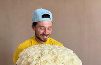 Vase filled with white roses held by smiling man wearing baseball cap.