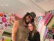 News Room : Game Of Thrones stars Jason Momoa and Emilia Clarke reunite 15 years after starring on iconic HBO series Huge man with curly hair, sunglasses, and a hat standing next to a smiling woman in black, vibrant colorful background.