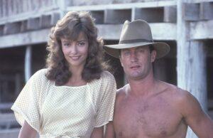 Young woman and shirtless man standing outdoors on wooden structure in vintage attire.