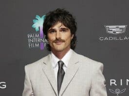 Stylish man in a light gray suit at a film festival event with Palm Springs International Film Festival logo in the background.