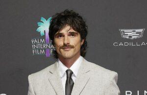 Stylish man in a light gray suit at a film festival event with Palm Springs International Film Festival logo in the background.