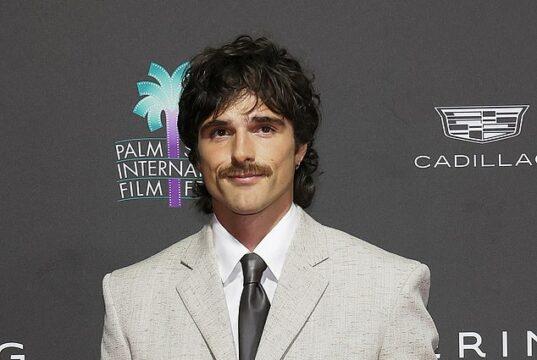 Stylish man in a light gray suit at a film festival event with Palm Springs International Film Festival logo in the background.