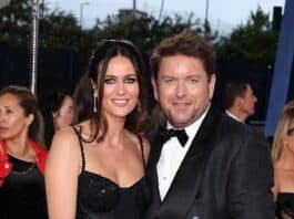 Elegant couple in formal black attire attending a glamorous event, surrounded by other guests.