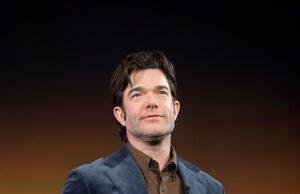 News Room : John Mulaney postpones his comedy shows in ‘unsafe’ Minneapolis after ICE killed mom-of-three Man in navy blazer on stage at a conference or event in front of a colorful background.