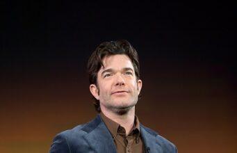 News Room : John Mulaney postpones his comedy shows in ‘unsafe’ Minneapolis after ICE killed mom-of-three Man in navy blazer on stage at a conference or event in front of a colorful background.