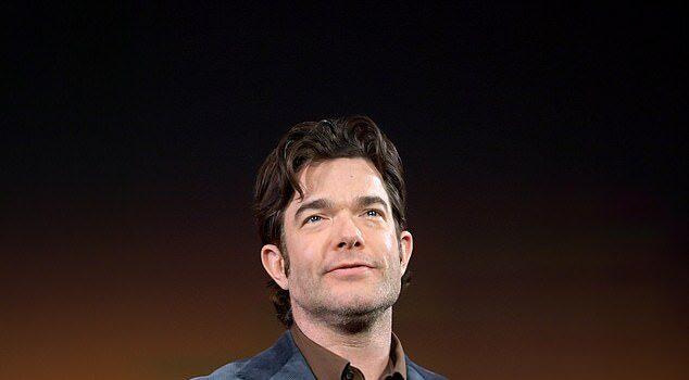 News Room : John Mulaney postpones his comedy shows in ‘unsafe’ Minneapolis after ICE killed mom-of-three Man in navy blazer on stage at a conference or event in front of a colorful background.