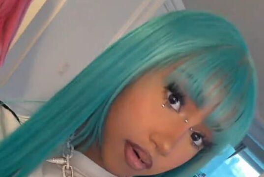 Vibrant turquoise-haired woman with jewelry and bold makeup in a modern room setting.