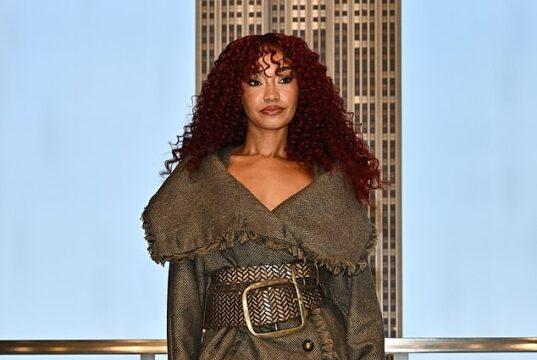 Stylish woman in a textured coat, patterned tights, and fur boots on city rooftop balcony at sunset.