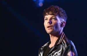 Young man performing on stage with microphone, dark background, casual stylish attire, tattoos on arm.