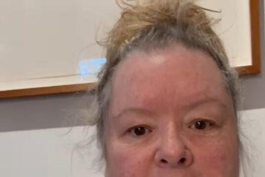 Uncertain woman with messy hair in casual grey shirt indoors, plain wall background.