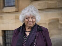 Senior woman with curly gray hair smiling, standing outdoors in front of historic building, wearing purple jacket and scarf.