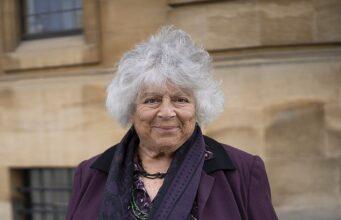 Senior woman with curly gray hair smiling, standing outdoors in front of historic building, wearing purple jacket and scarf.