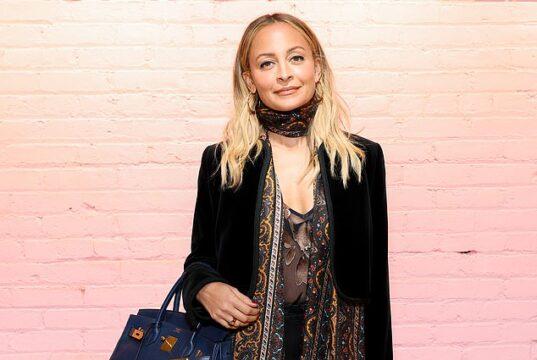 Fashionable woman with blonde hair posing against a pink brick wall, holding a blue handbag.