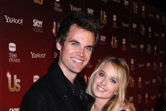 Smiling young couple at entertainment event, standing close together in front of media backdrop.