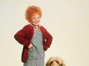 Adorable young girl with curly red hair in a vintage dress and cardigan standing next to a fluffy, friendly mixed breed dog.