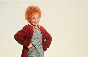 Adorable young girl with curly red hair in a vintage dress and cardigan standing next to a fluffy, friendly mixed breed dog.