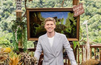 News Room : PETA’s scathing letter to Robert Irwin ahead of his stint on the upcoming season of I’m A Celebrity… Get Me Out Of Here A young man in a light gray suit smiling at a desk decorated with wildlife sculptures outdoors.