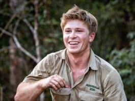 Young man smiling outdoors in khaki shirt at the zoo, holding a small item in his hand.