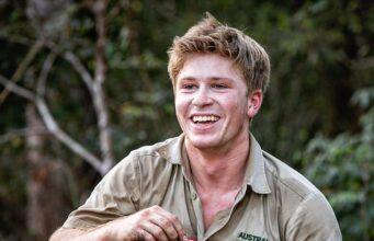 Young man smiling outdoors in khaki shirt at the zoo, holding a small item in his hand.