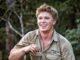 News Room : Robert Irwin caught off guard after radio host asks personal question Young man smiling outdoors in khaki shirt at the zoo, holding a small item in his hand.