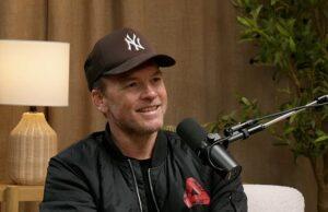 Young man with a microphone, wearing a New York Yankees cap, during podcast interview indoor setting with wooden decor and plants, smiling confidently.