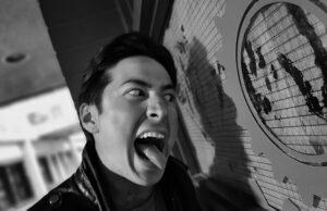 Young man making a funny face, sticking out his tongue near a large clock on a wall.