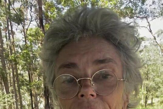 Elderly woman with glasses and floral blouse outdoors in sunny forest setting.